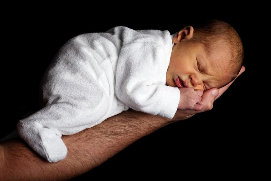 A small baby in a white sleepsuit sleeps in a man's hand and forearm.