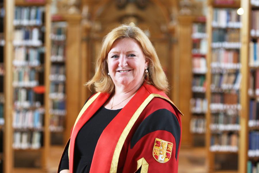 Woman with shoulder length blonde hair smiles at the camera. In the background are the out of focus bookshelves of Bangor University library.
