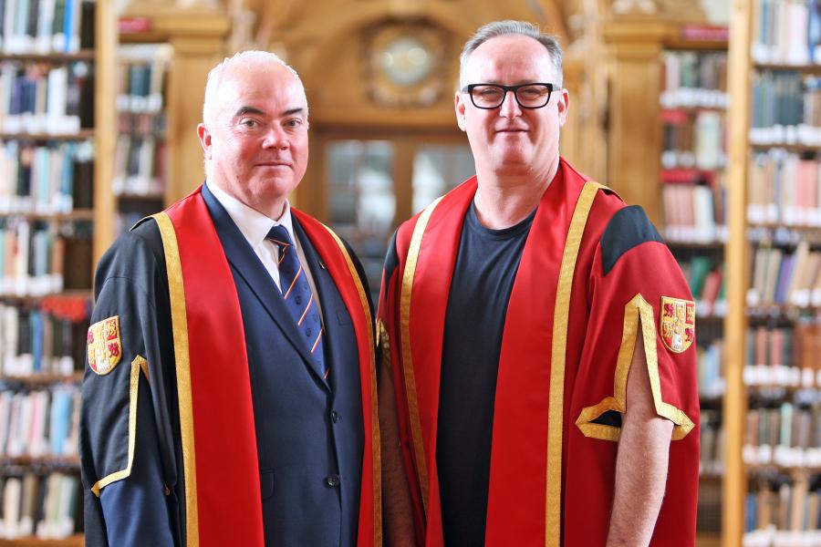 Two moddle aged men in academic gowns- Sasha on th eright has a red gown over a t shirt