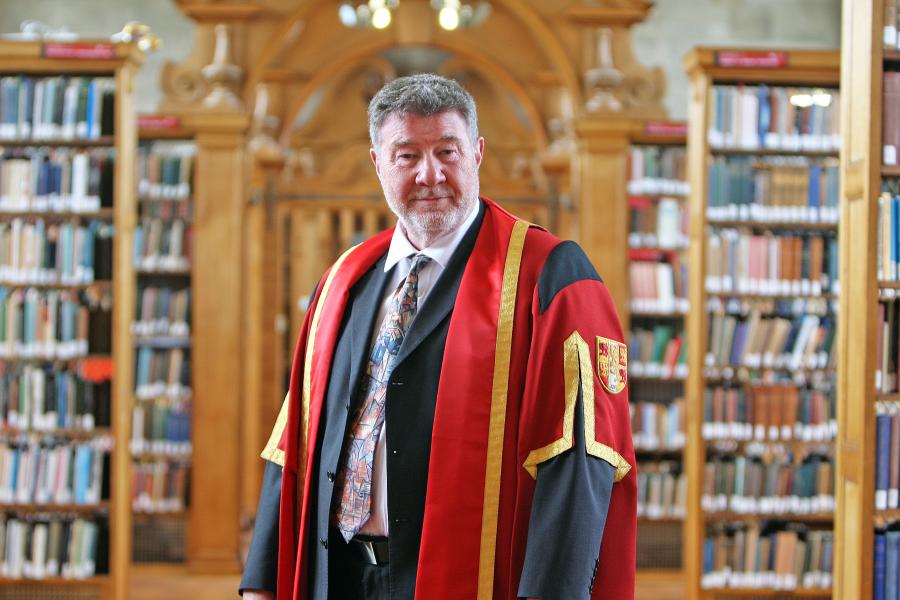 Man in  suit nd flowery tie and a red graduation gown stands in Bangor University library