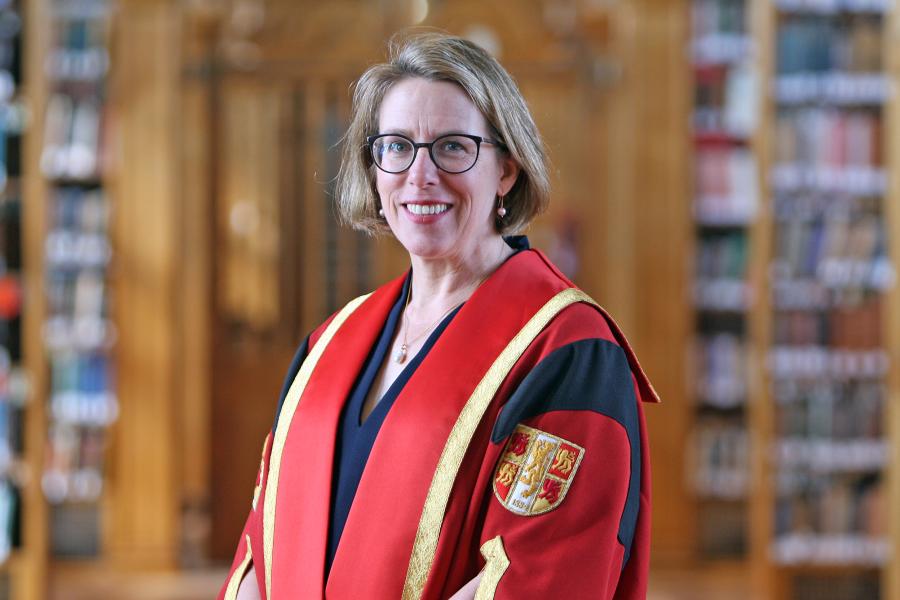 Woman with shoulder-lengths blonde hair and large glasses smiles to camera. She is wearing a red graduation gown and standing in front of out of focus book shelves.