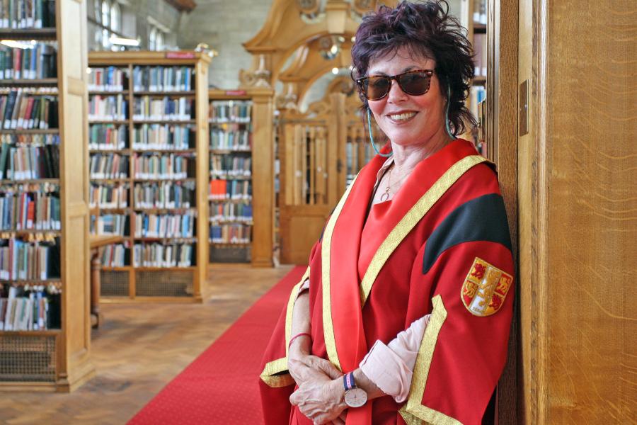 Ruby wax in red academic gown and dark glasses, leans on the end of a book shelf in the Shankland Library at Bangor University