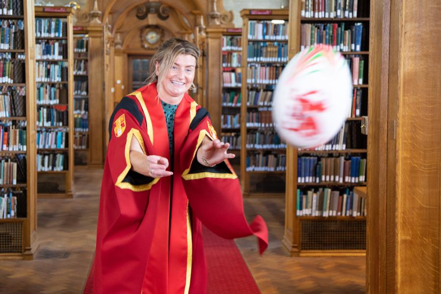 Young wonam in red graduation gown moves to catch a rugby ball spinning in the foreground