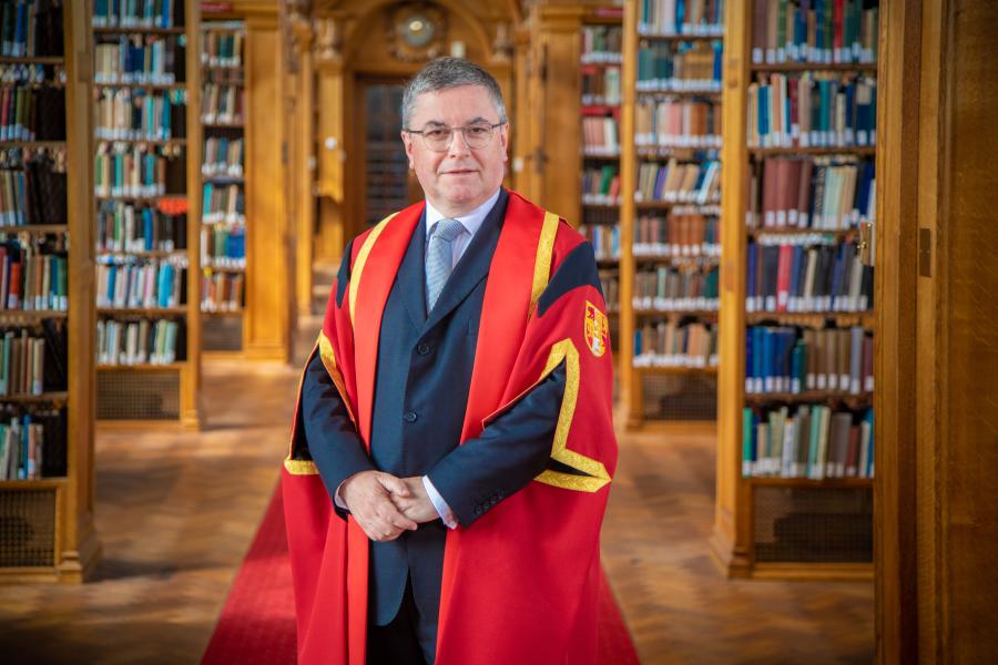 Man with short grey hair and spectacles stands in red graduation robe in Bangor University's library 