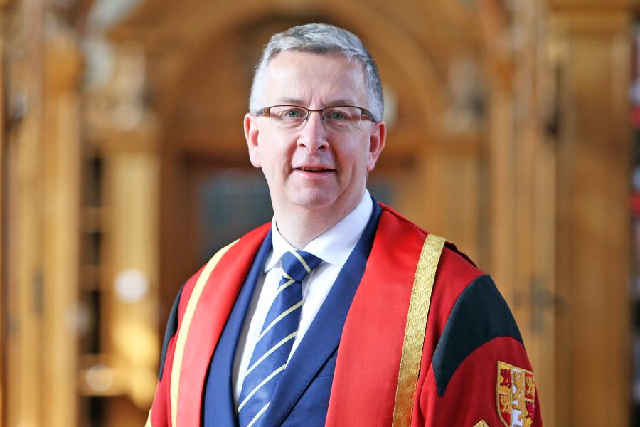 Grey haired man with glasses and blue suit and blue striped tie wears a red graduation gown in Bangor University library.