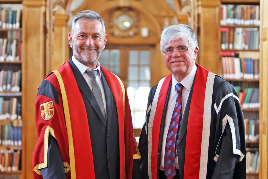 Two men in academic dress, the one on the left has a red gown. they stand in a library.