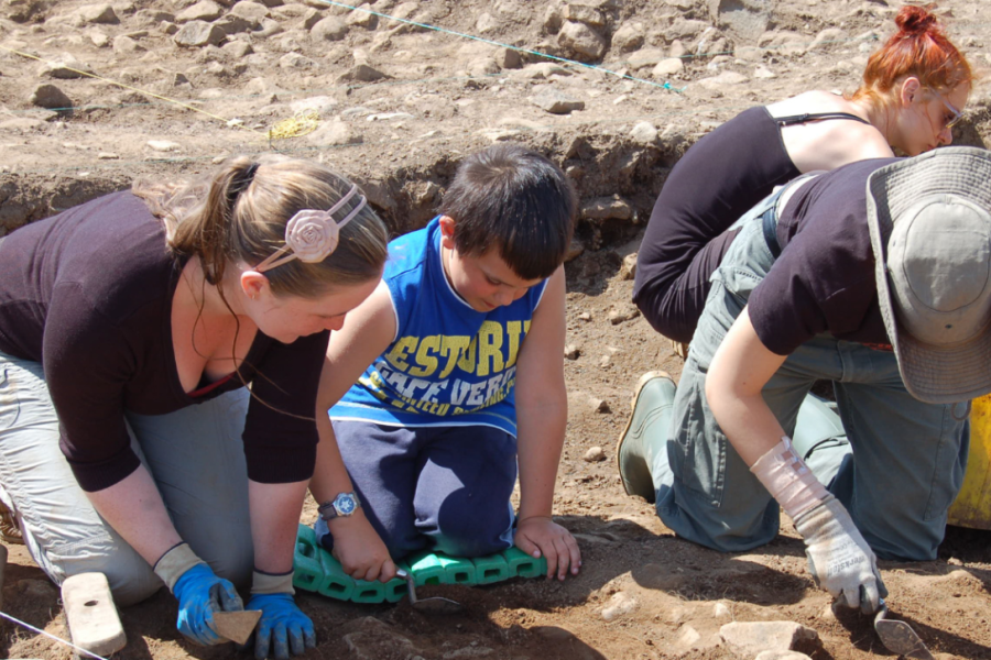 Adults and children kneel on their knees on the ground, taking part in a community archaeological dig.