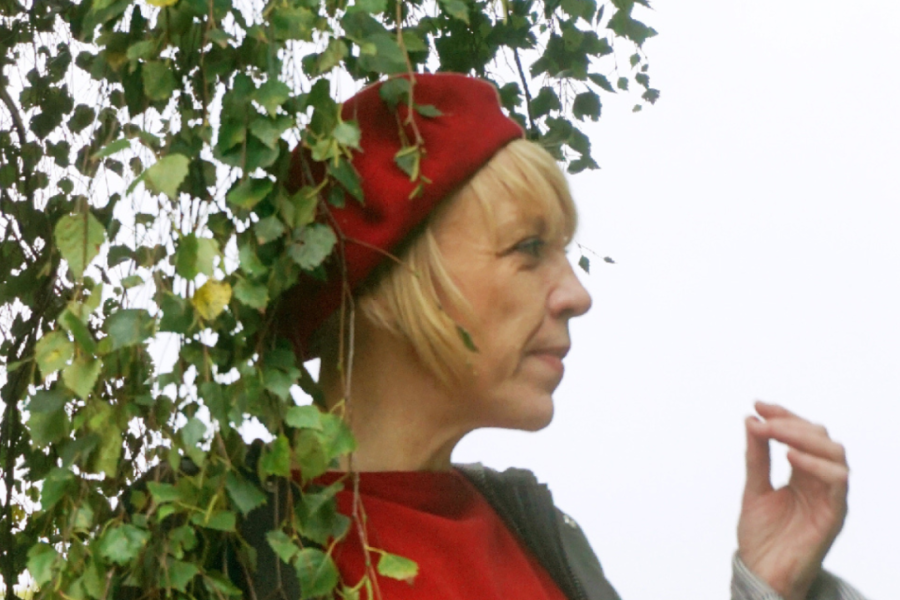 Woman with blonde hair wearing red and a red beret stands directly under a tree in leaf.