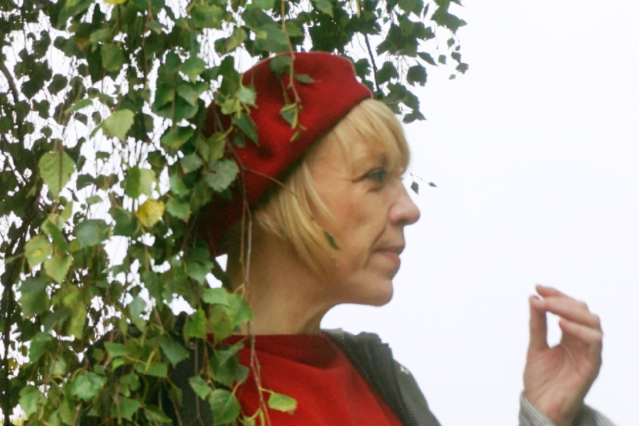 Merch yn gwisgo coch ac efo beret coch a gwallt melyn yn sefyll yn agos o dan dail coedn with blonde hair wearing red and a red beret stands directly under a tree in leaf.