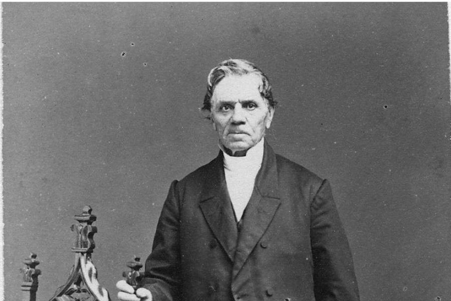 Hen ffotograff du a gwyn o'r Parchedig Robert Everett yn sefyll gyda'i law ar gefn cadair cerfiedig.hoto of Rev Roberts Everett  standing with his hand on the back of a carved chair