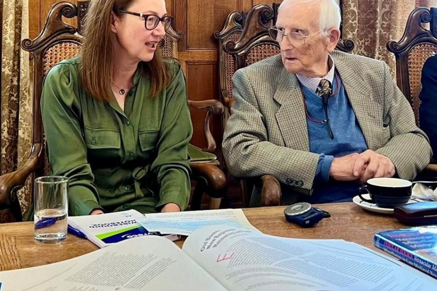  Woman in green shirt and straight brown hair is seated talking t o older man in suit.