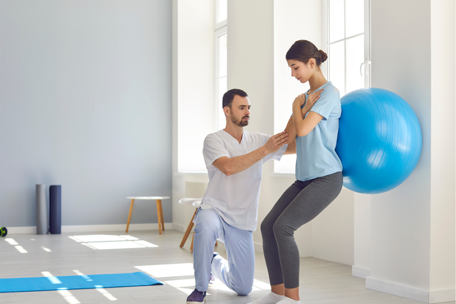 Female leaning on an exercise ball against a wall, with a male helping her