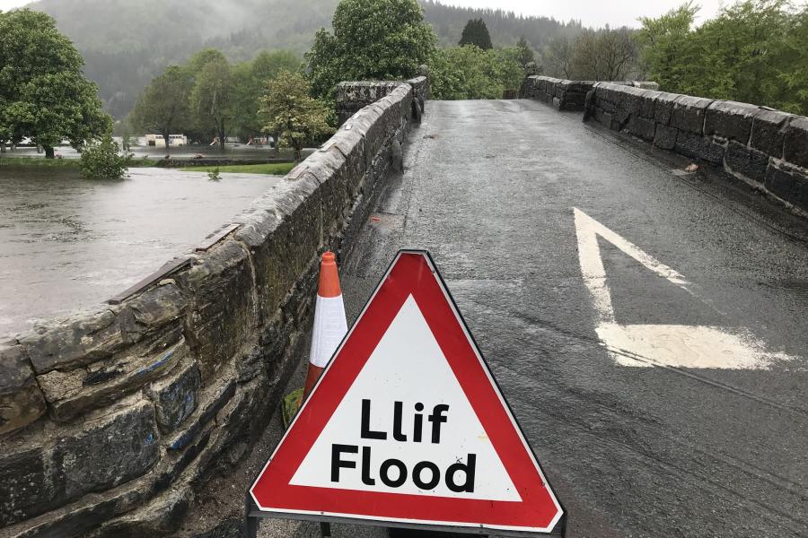 A Flood warning sign sits at the end of a bridge, beyond the river floods through trees.