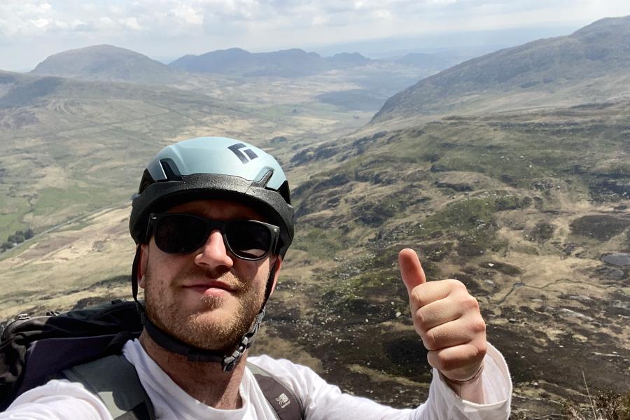 George,  yn gwisgo sbectol haul a helmed dringo yn rhoi bawd fyny wrth iddo hyfforddi ar gyfer yr alldaith Last Pole. Mae'n amlwg ar dir uchel ac mae mynyddoedd i'w gweld yn y cefndir.t gives a thumbs up sign,  as he prepares for his Last Pole expedition. There are mountains in the background.