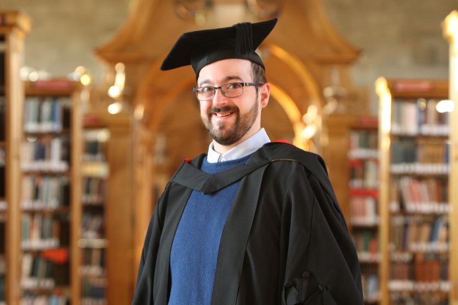 Man with academic gown blue jumper and spectacles stands in library