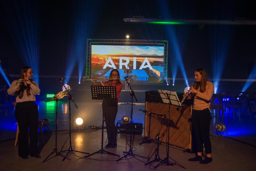 A woodwind trio performing in front of a screen which reads Aria
