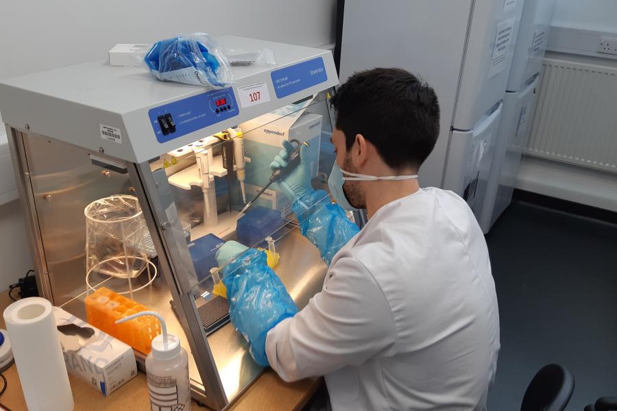 Person in protective clothing, wearing long blue gauntlets and a lab coat, testing in a fume cupboard.