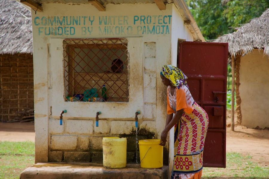 A woman fills a large yellow bucket from a water pimp. On the wall above her are the words Community water prooject funded by Mikoko Pamoja