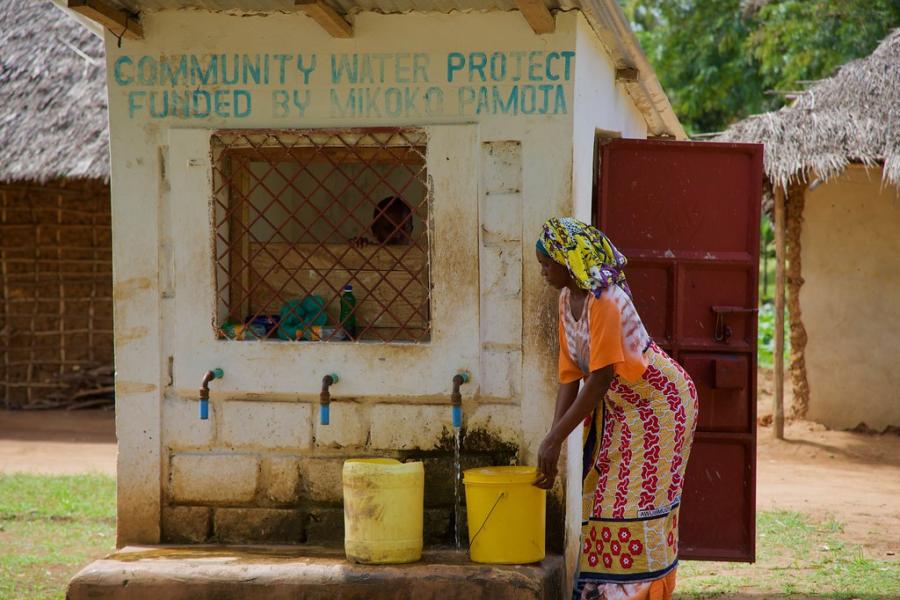 Merch y  lllenwi bwced mawr efo dŵr o bwmp. Ar y wal uwchben, mae'r geiriau Community water prooject funded by Mikoko Pamoja