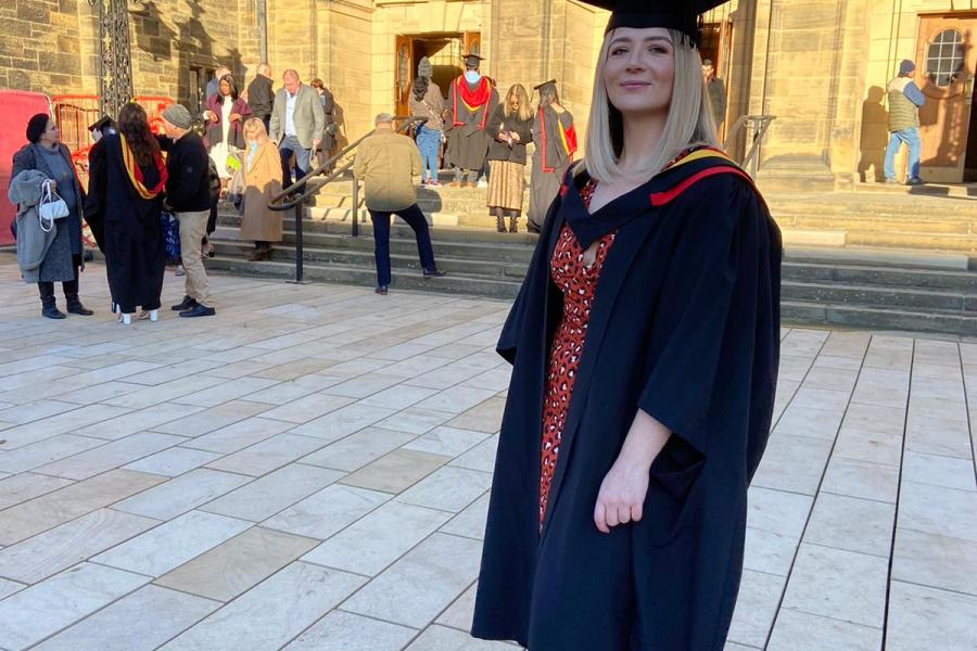 Elen (Ellie) Rankenhohn smiling to camera wearing her cap and gown at graduation outside the main arts building. 