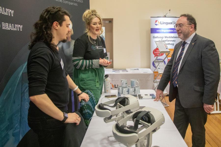 Two younger people chat across a table with Vice Chancellor in a suit. There are VR headsets in the foreground