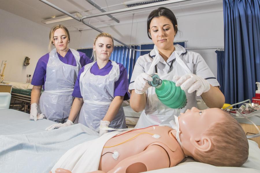 Dwy myfyrwraig nyrsio yn gwylio arddangosiad wrth ochr gwely  gyda dymi yn y gwely.ents at a bedside watching someone with a dummy