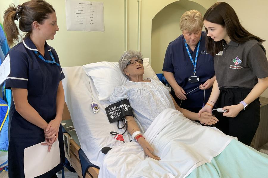 YOung girl approached a dummy in a bed- there are nurses on either side.