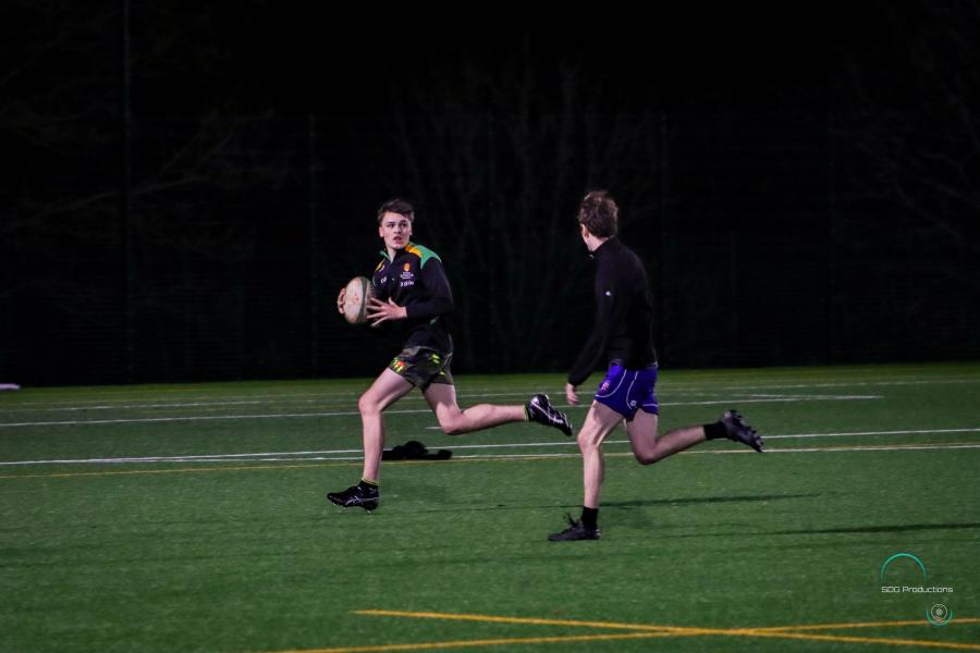 Two rugby players, one running with the ball looks back towards the other approaching from the side.