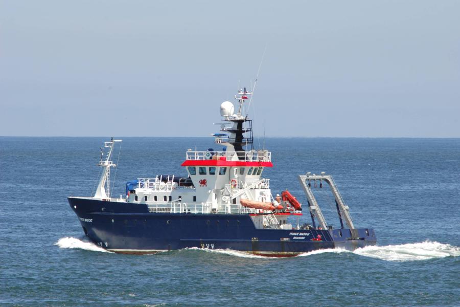 A blue and white research vessel creates a bow wave in a blue sea