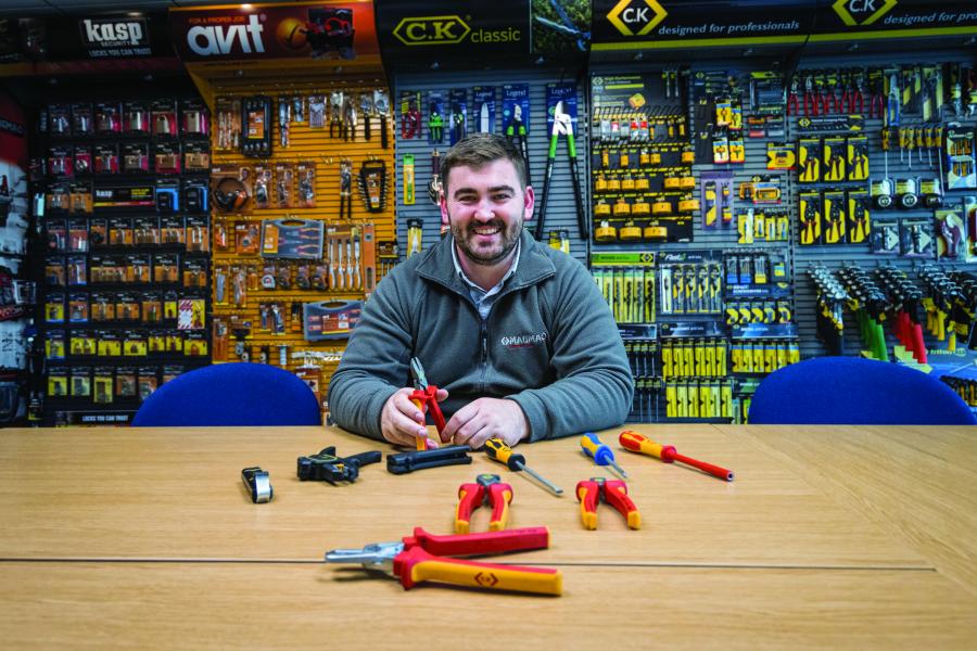 Mathew Jones, Product Design graduate sitting in a tools shop with tools laid out on a table in front of him and shelves housing tools behind him