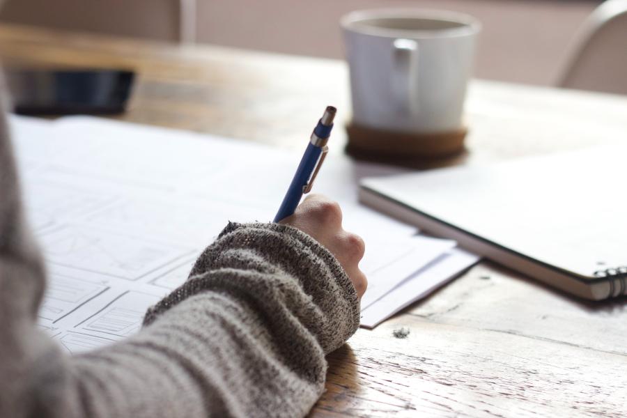 person writing on brown wooden table with a cup in the backgroun