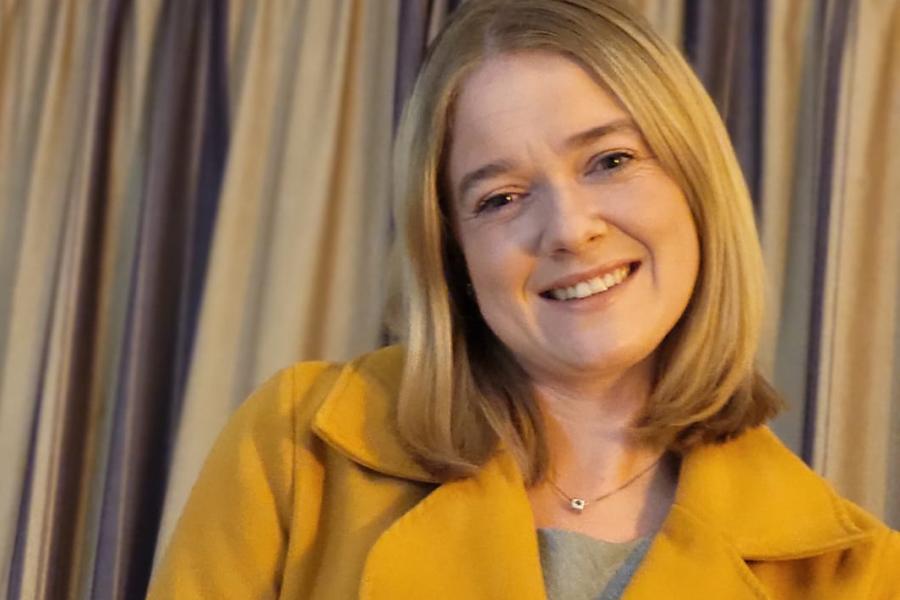 Megan Phillips smiling to camera in orange coat with curtain behind her