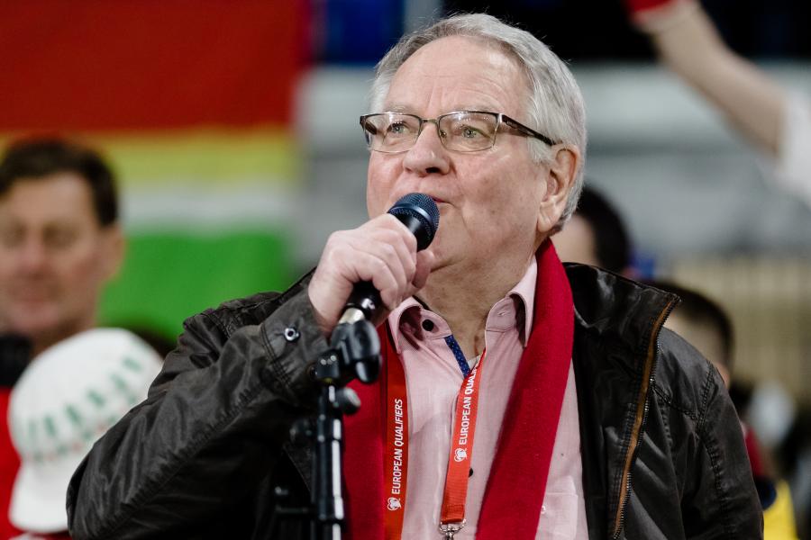 Dafydd Iwan holds a microphone to his mouth in an outdoor football setting.