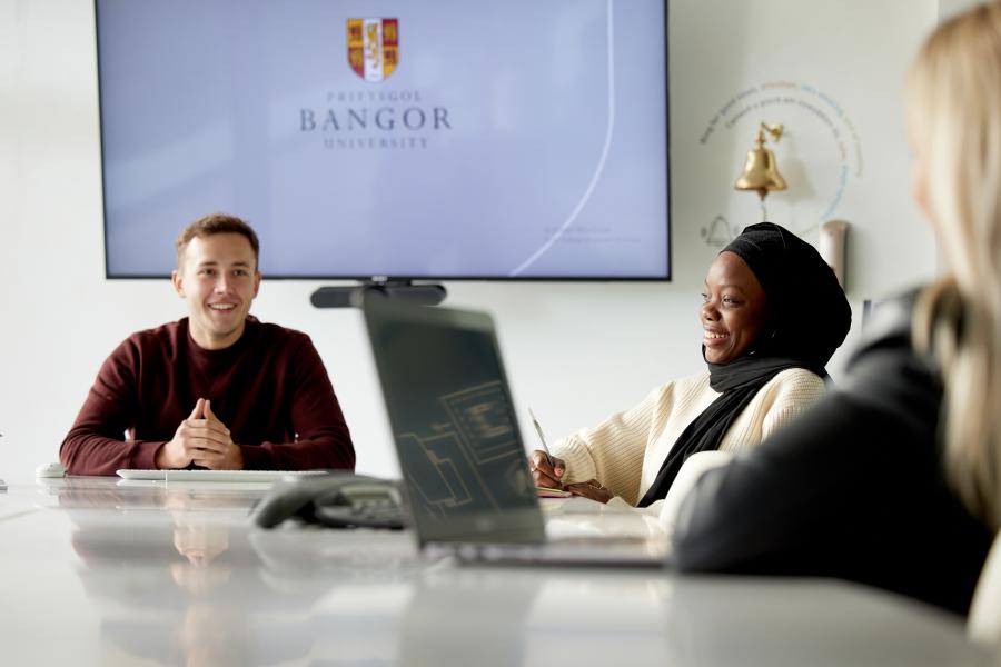 Students in MSparc smiling. There is a laptop on the table and Bangor University logo is in the big screen in the background. 