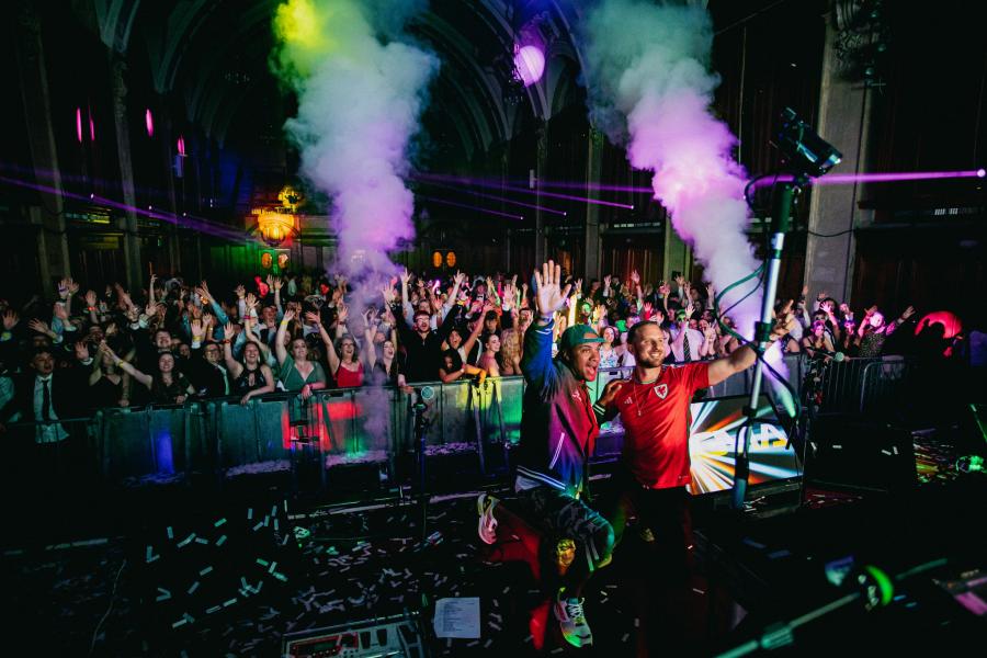 The inside of Academi student night club with two DJs and room full of people cheering. Smoke and colour-full lights. 