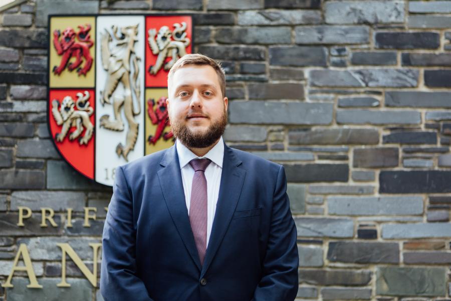 Bearded man in bllue suit in front of the Bangor University shield