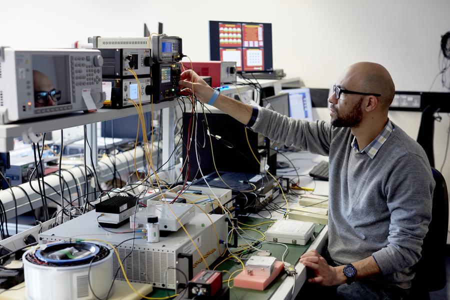 Engineering student sitting at a desk, looking to the left, with one arm raised to press buttons on an item of electrical equipment.