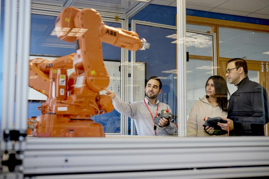 Two students and one lecturer standing in a laboratory, looking to the left, one pointing at an orange robotic arm.