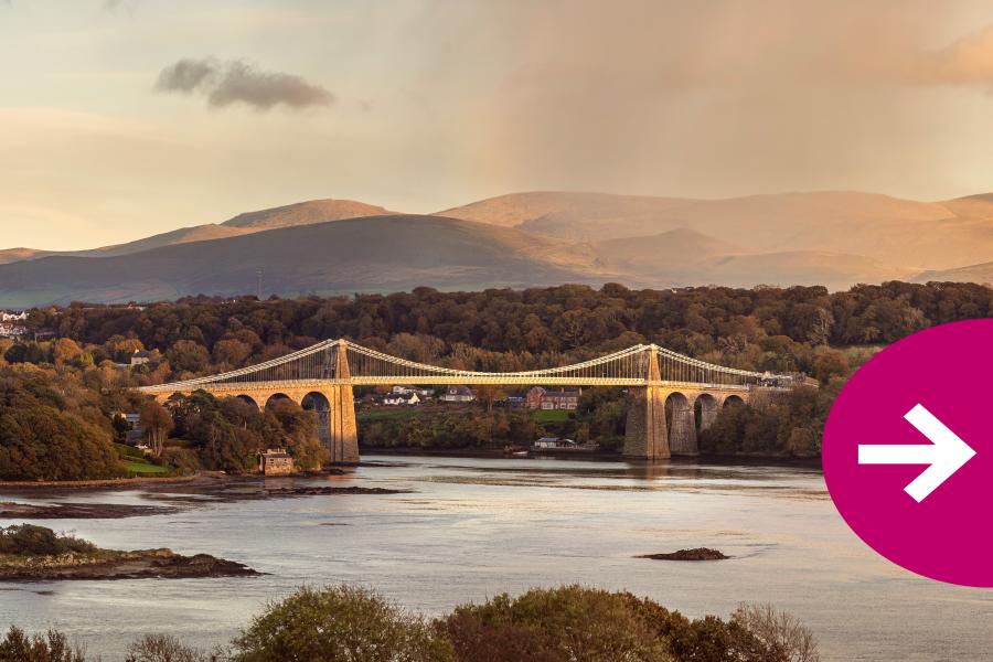 Llun o Bont Menai uwch ben Y Fenai gyda mynyddoedd Eryri yn y cefndir. Mae'r llun yn cynnwys saeth i'r dde i annog clicio drwodd i'r llun nesaf.