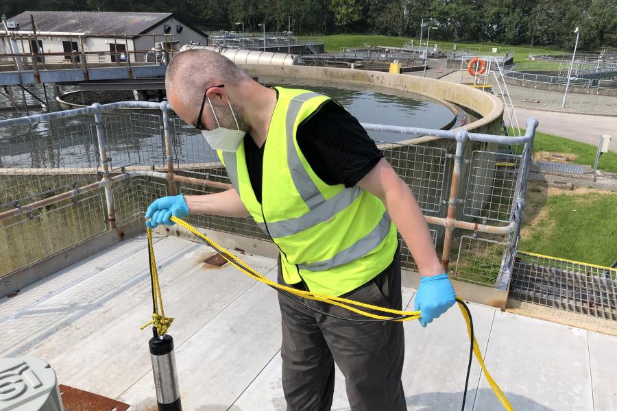 A researcher wearing a high viz vest, facemask and blue plastic gloves, taking samples for viral and plastics analysis at an urban wastewater treatment plant