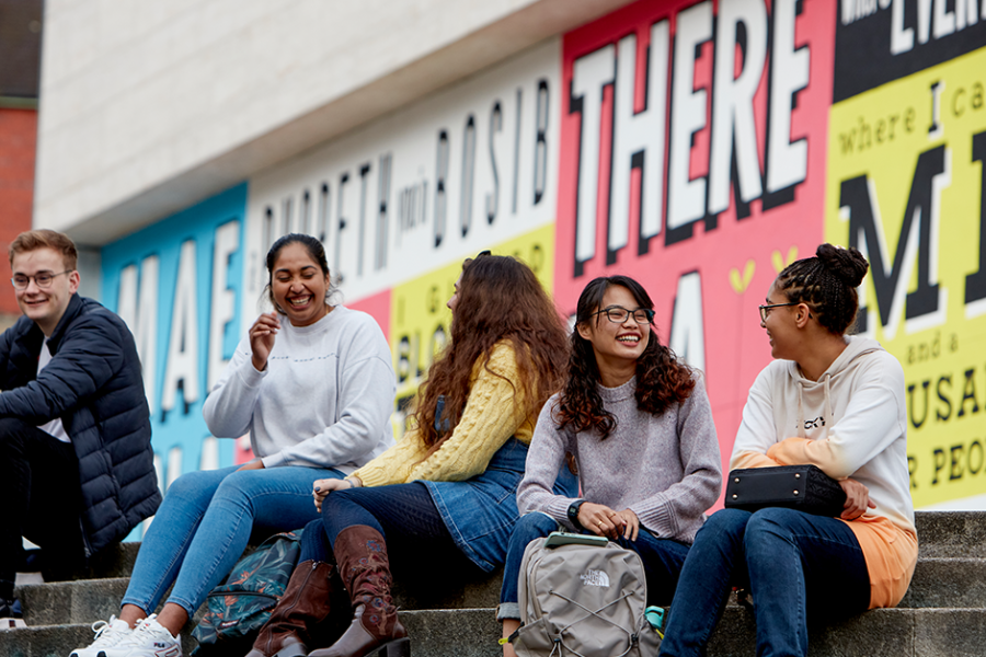 students sitting outside Pontio on the steps in front of a colourful mural.