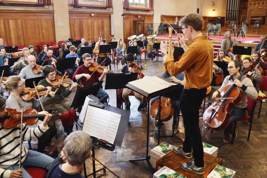  A young man in mustard coloured shirt conducts a n orchestra of adults and young people