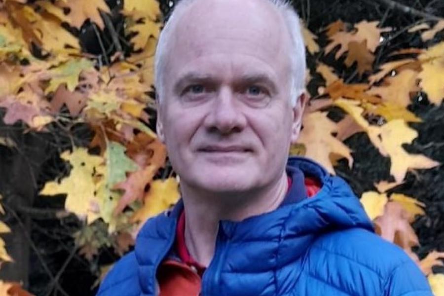 Head and shoulders of man in blue coat with background of leaves.