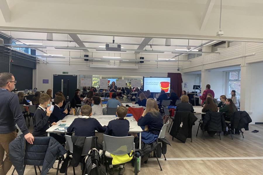 A University room filled with round tables of school children attending a Youth Sumit, lecturer is standing at the front of the room next to a power point screen delivering a presentation 