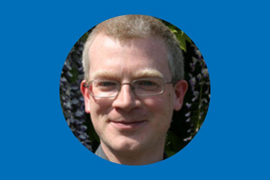 Bespectacled man with shorth blond hair, head in blue circle border