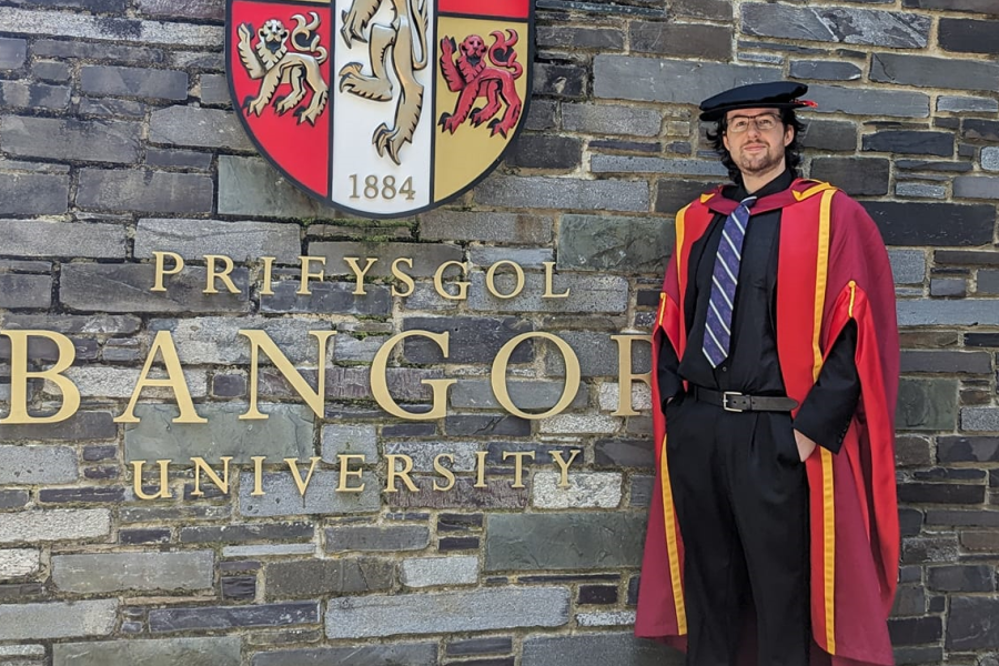 Dr Alex Baxendale in graduation ceremony robes standing outside main arts in front of the Bangor University emblem on the wall