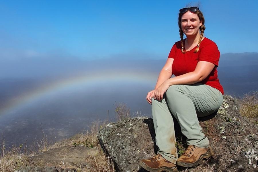 Dr Awel Vaughan-Evans yn eistedd ar graig gydag awyr las ac enfys yn y cefndir.