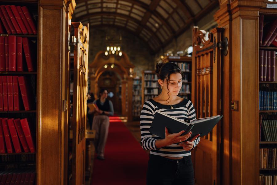 A student stands in the central aisle of Shankland Library reading a book, surrounded by bookshelves with warm light filtering through high, arched windows. The vaulted ceiling and chandeliers add a sense of quiet grandeur.