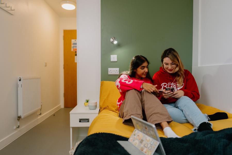 Two students sitting on a bed looking at a moblie phone in a room in St Mary's halls.