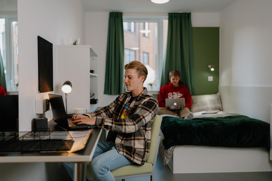 Two students in a room in St Mary's halls. One is reading on the bed, another is sitting at a desk working on a laptop